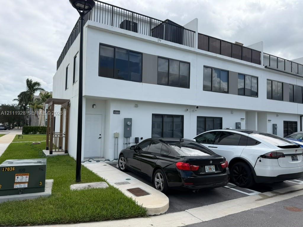 Modern two-story townhouse exterior featuring a smooth stucco facade, expansive rooftop terrace with black metal railing, large rectangular windows, and a paved private parking area