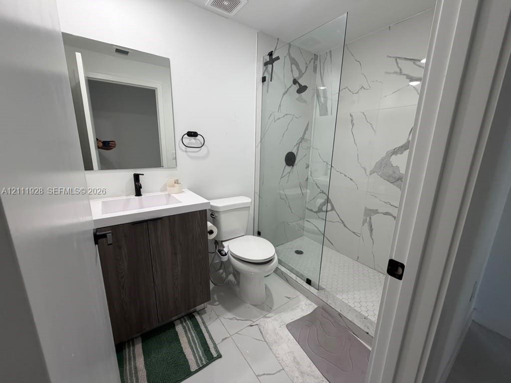 Modern bathroom featuring a wood-finish vanity, white countertop with integrated sink, walk-in shower with floor-to-ceiling veined tile, glass enclosure, and matte black fixtures