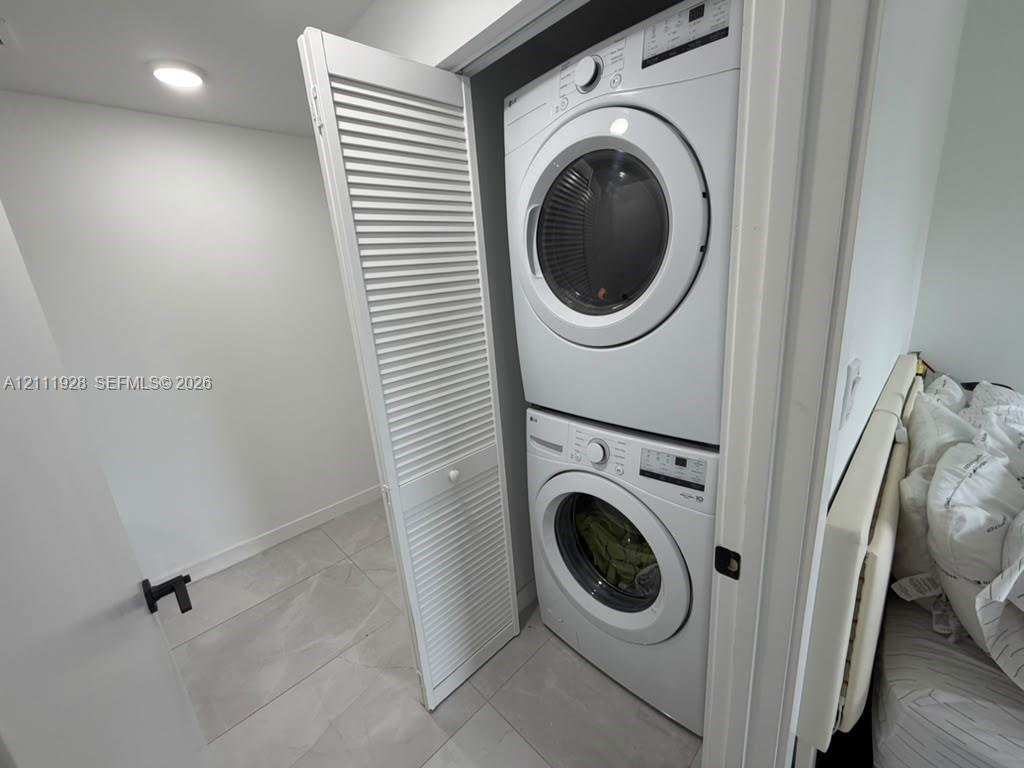 Stacked white front-load washer and dryer unit enclosed by white louvered bifold doors, featuring tile flooring and recessed ceiling lighting