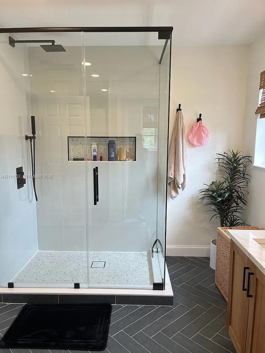 Walk-in shower featuring a glass enclosure with matte black hardware, a rainfall showerhead, a handheld sprayer, and a recessed tiled niche, complemented by charcoal-toned herringbone floor tiling