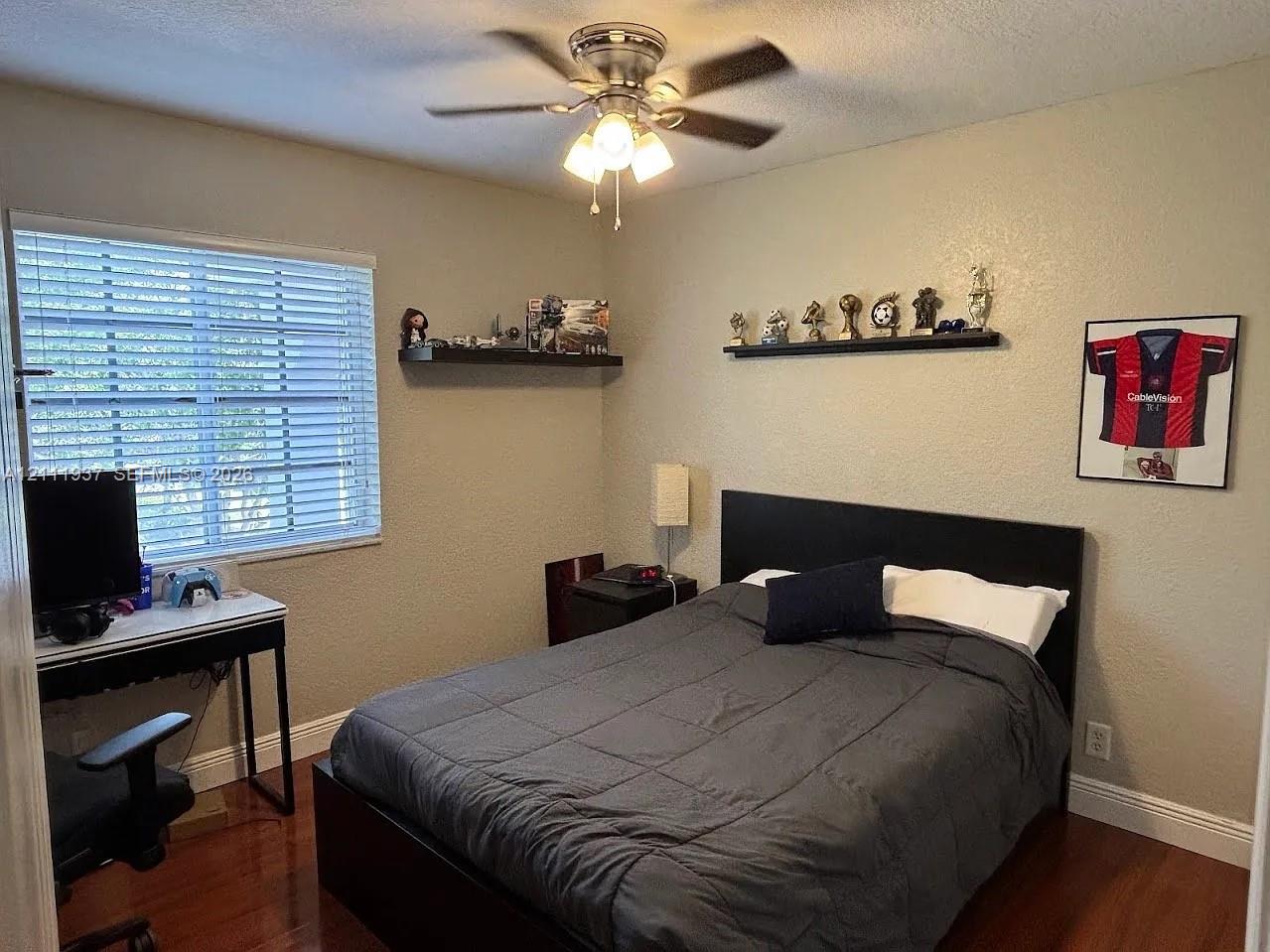 Wood-finish flooring, ceiling fan with integrated light fixture, two mounted wall shelves, and windows featuring horizontal blinds