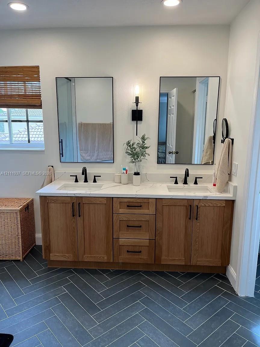 Double vanity with a stone countertop, under-mount sinks, wood cabinetry, matte black hardware, and herringbone-patterned tile flooring