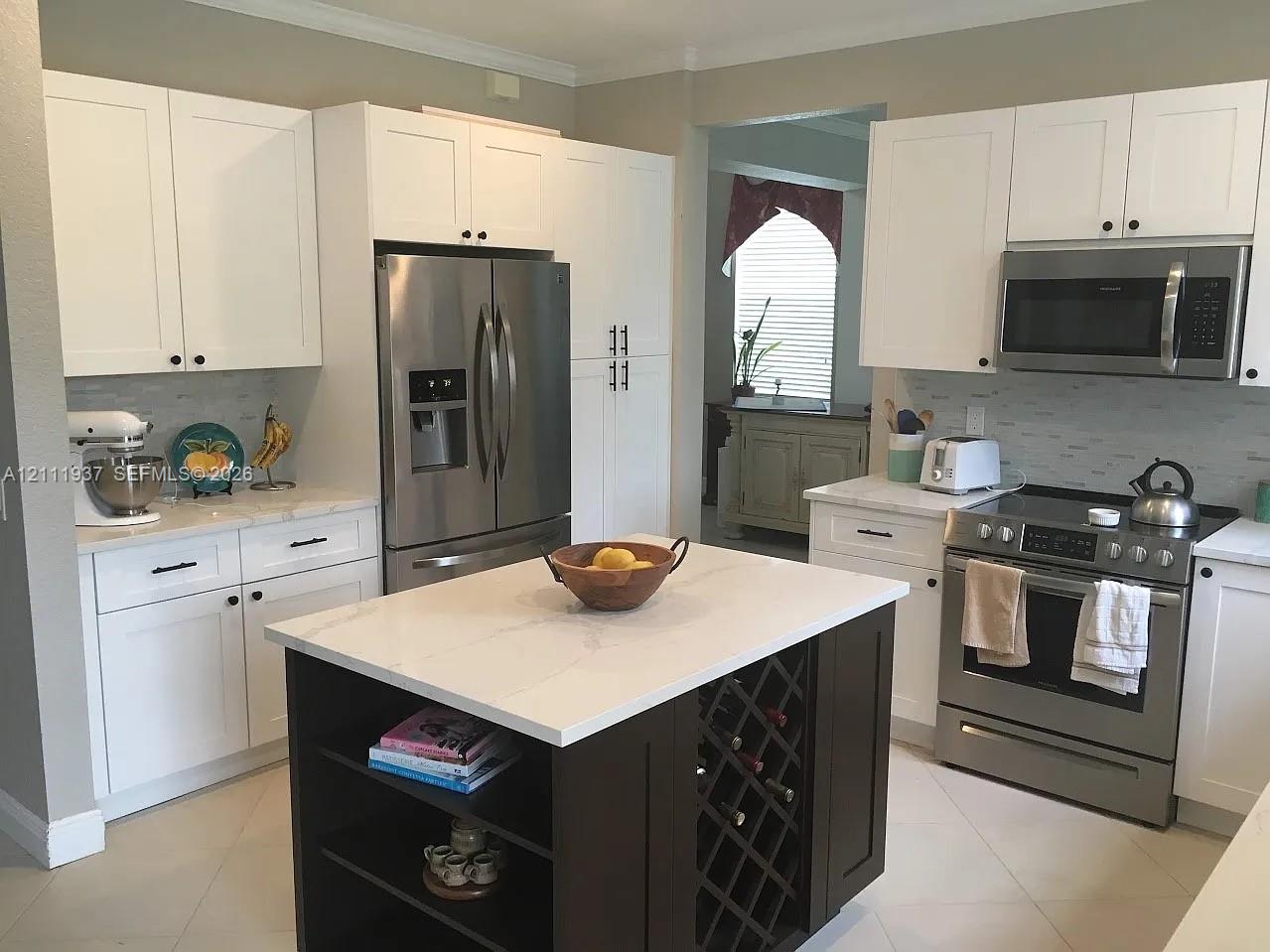 Kitchen featuring white shaker-style cabinetry, a central island with built-in wine storage and open shelving, light-toned stone-finish countertops, stainless steel appliances, and a tiled backsplash