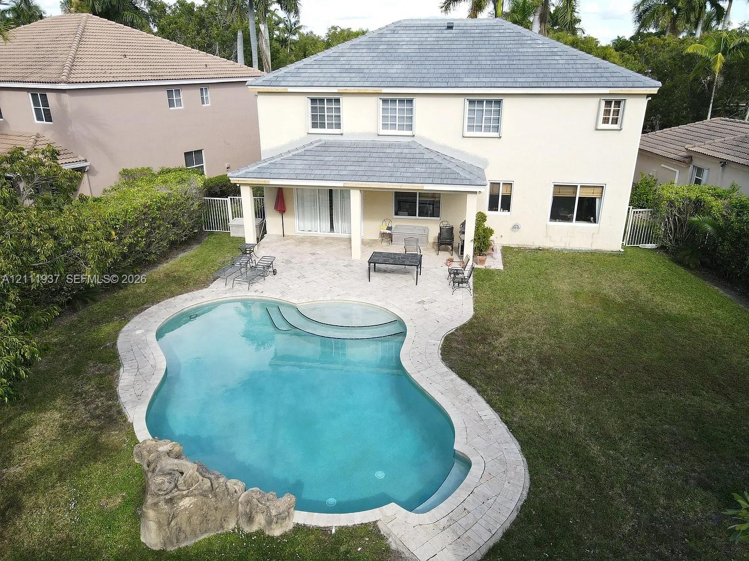 Rear exterior featuring a covered patio, paved pool deck, freeform swimming pool with integrated rock feature, and a manicured lawn