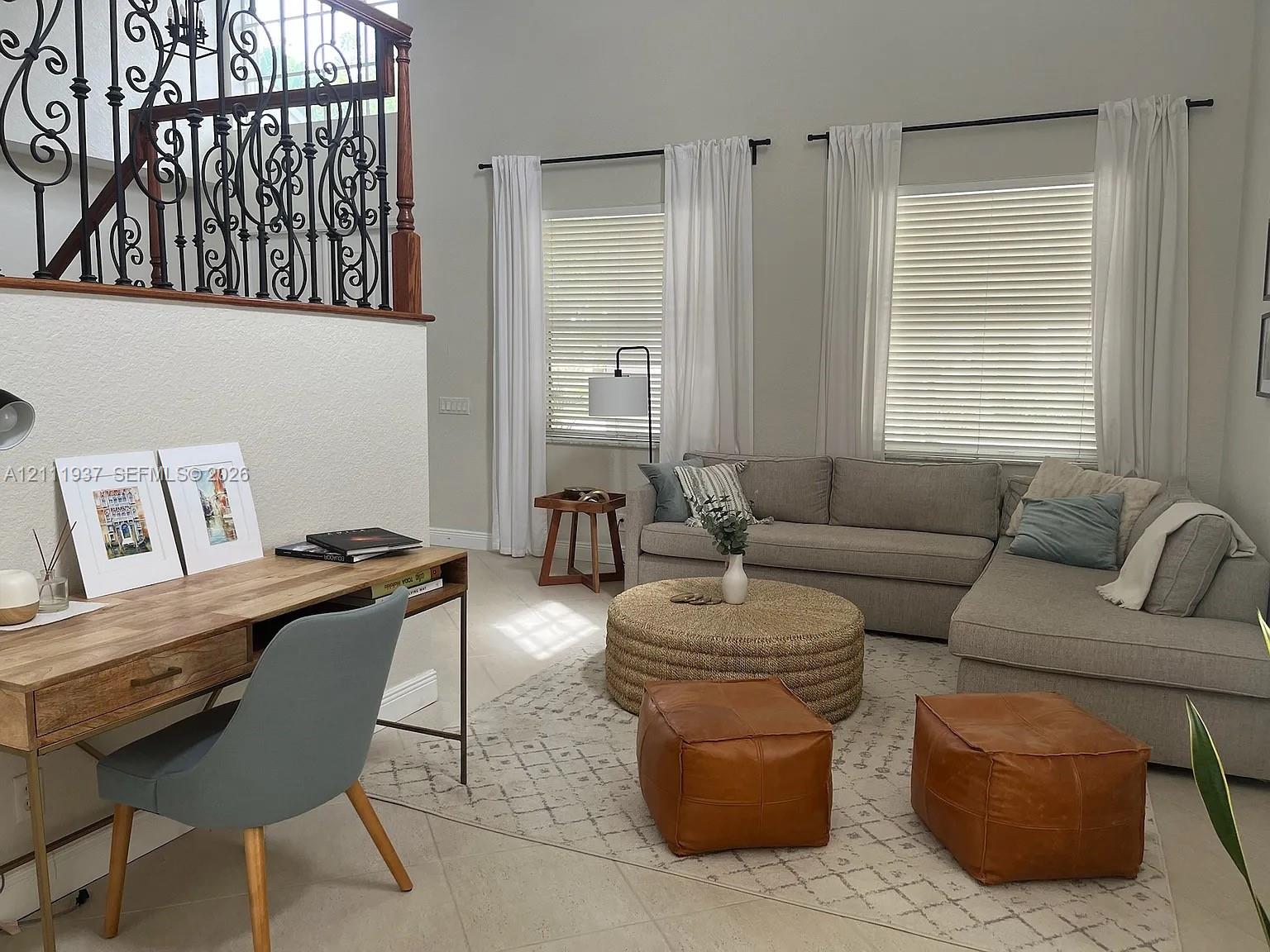 Double-height ceilings, wrought iron staircase railing with a wood handrail, neutral ceramic tile flooring, and large windows with horizontal blinds