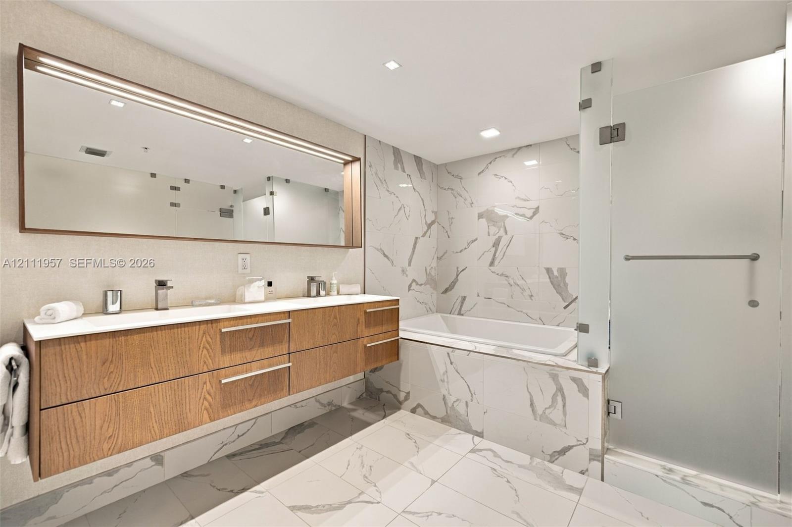 Floating wood-finish vanity with dual sinks, large integrated mirror, walk-in shower with frosted glass enclosure, and stone-tile flooring and wall surrounds