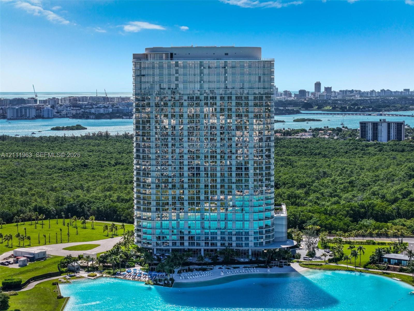 High-rise architectural exterior featuring floor-to-ceiling glass paneling, wraparound balconies, and a ground-level turquoise recreational lagoon