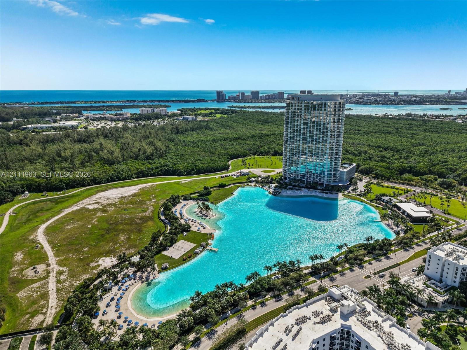 Aerial perspective featuring a high-rise residential structure, a expansive turquoise lagoon, sand shoreline, and lush surrounding greenery