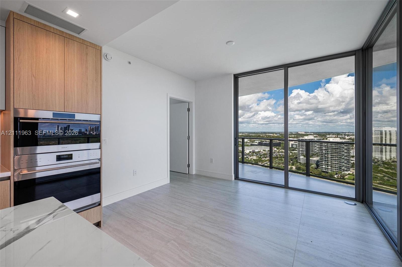 Modern kitchen featuring wood-finish cabinetry, a waterfall-edge stone countertop, built-in stainless steel appliances, floor-to-ceiling glass panel walls, and large-format tile flooring
