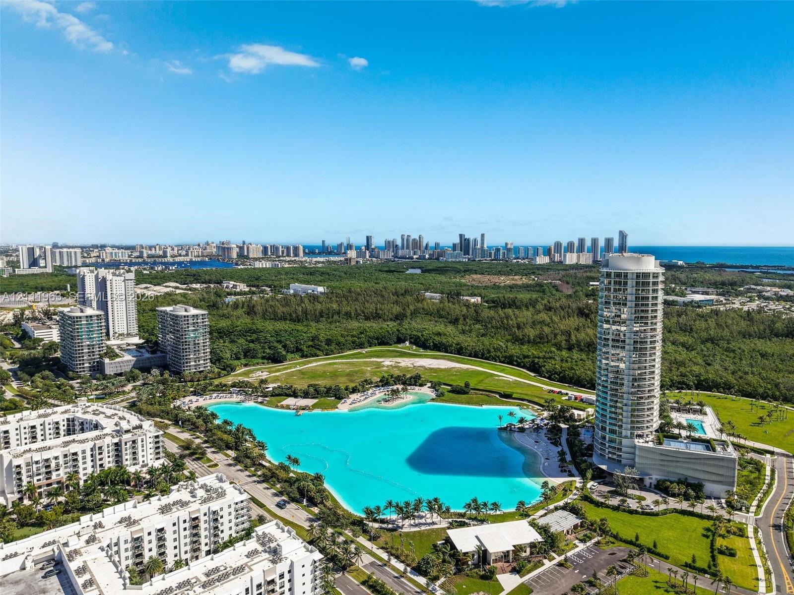 Aerial perspective of a high-rise residential tower featuring an on-site swimming pool, expansive turquoise lagoon, landscaped green spaces, and parking facilities