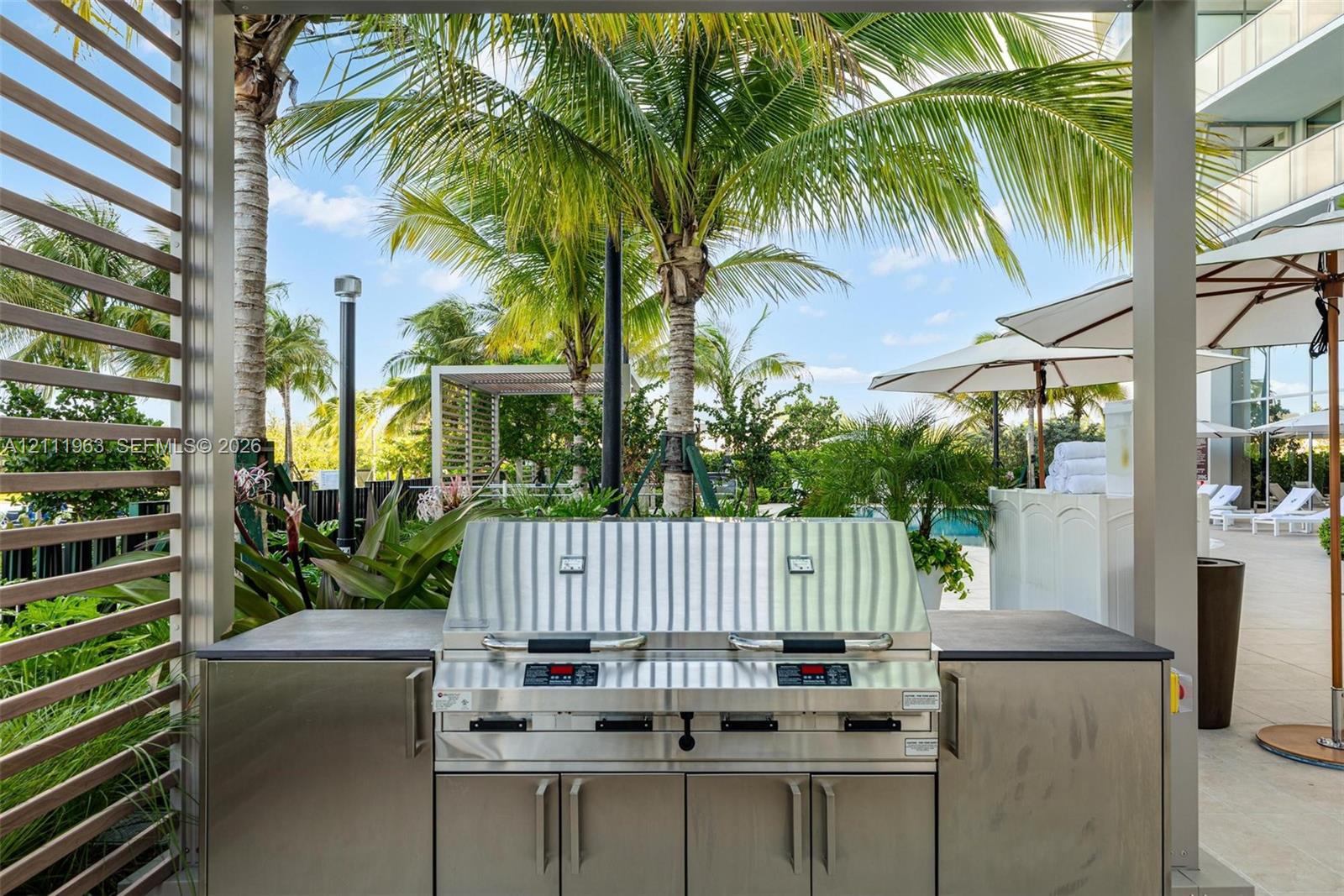 Expansive stone-paved patio featuring a covered pergola, built-in outdoor kitchen, integrated landscape lighting, and mature palm trees