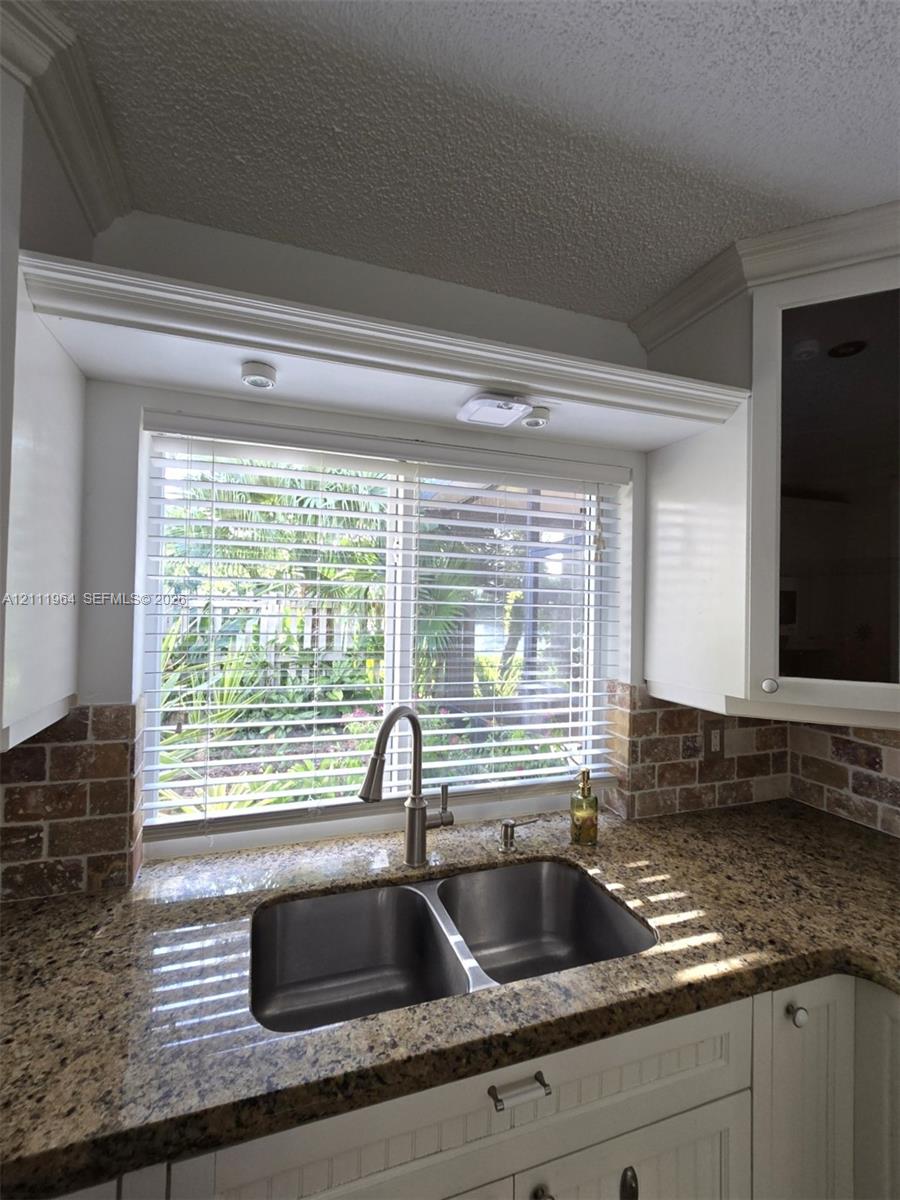 Kitchen area featuring white cabinetry with crown molding, stone-finish countertops, brick-style tile backsplash, and a double-basin stainless steel sink with a high-arch faucet