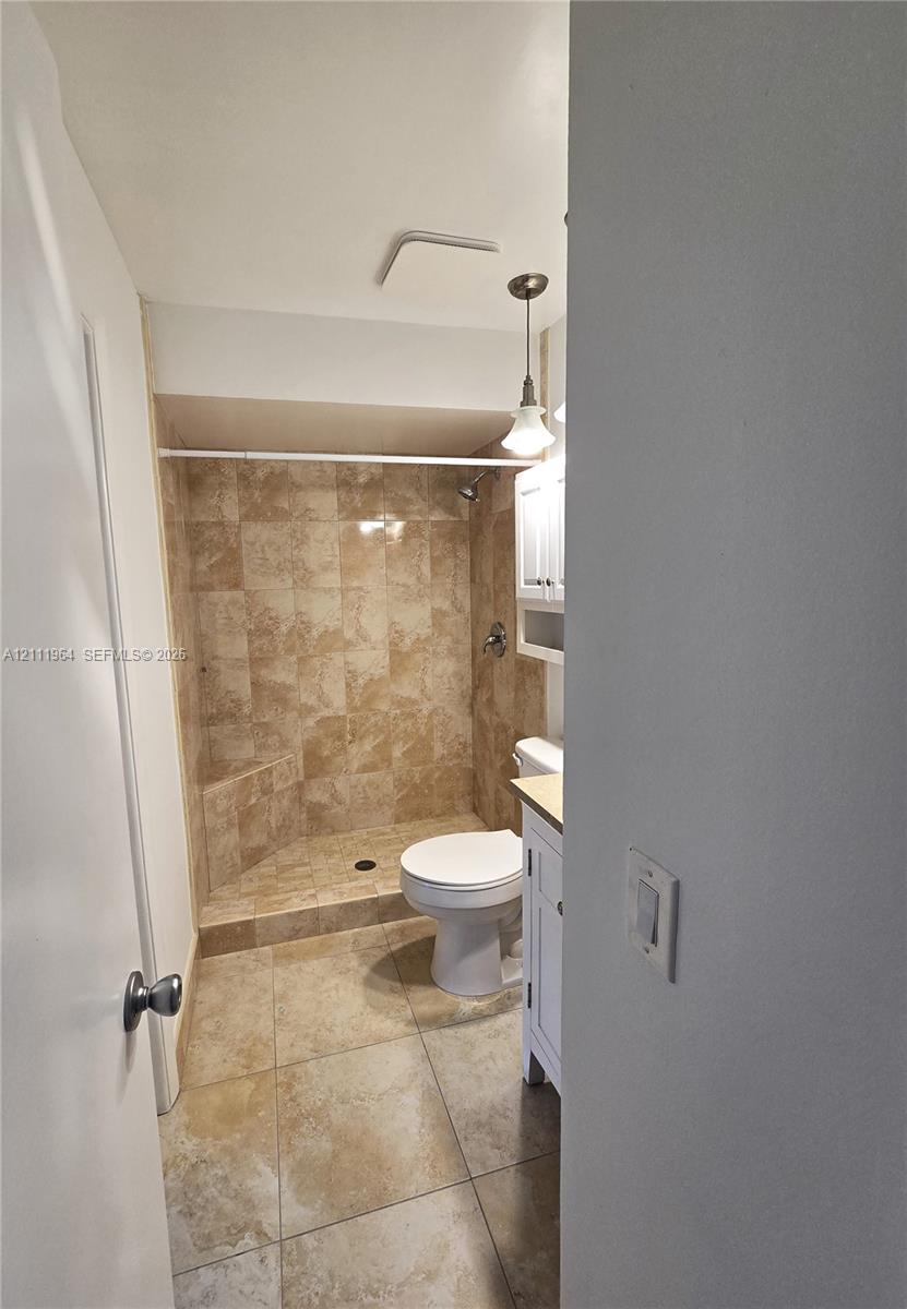 Walk-in shower with stone-tile surround, neutral ceramic tile flooring, white vanity cabinet, and pendant lighting