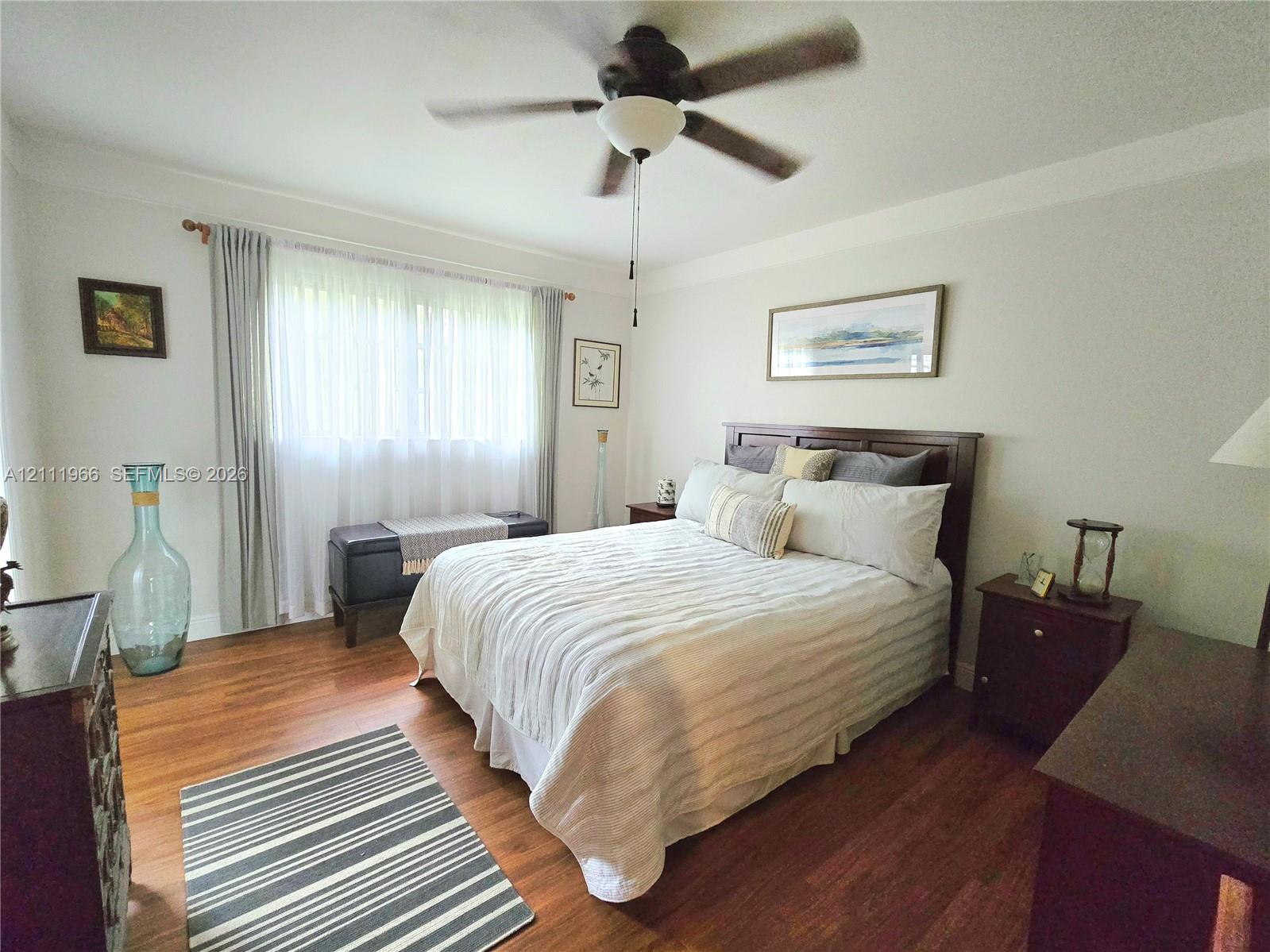 Wood-finish flooring, multi-pane window with neutral drapery, and a ceiling-mounted fan with integrated lighting