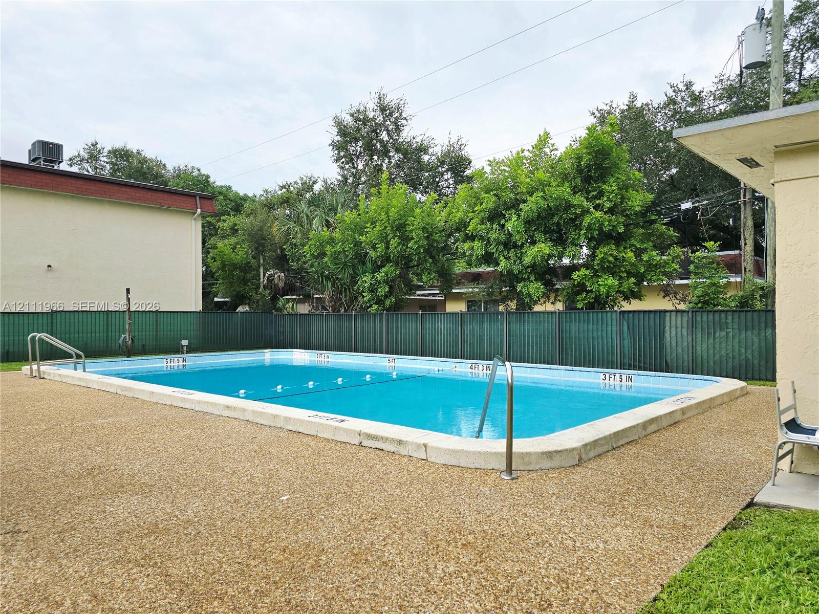 Rectangular in-ground swimming pool featuring a pebble-textured deck, metal handrails, and a full-height privacy fence