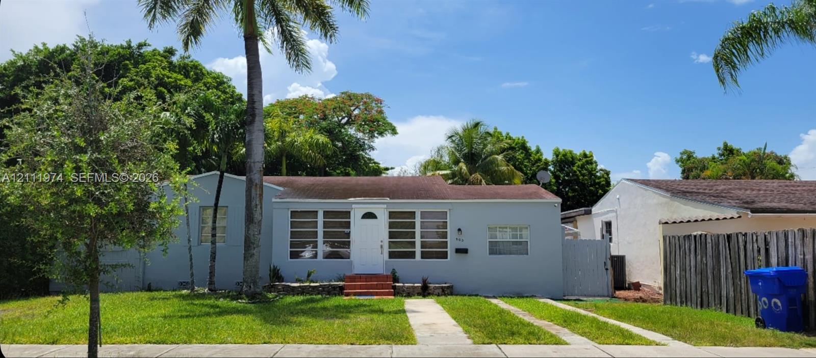 Single-story exterior featuring a light gray facade, multi-pane windows, a concrete walkway, and mature palm trees