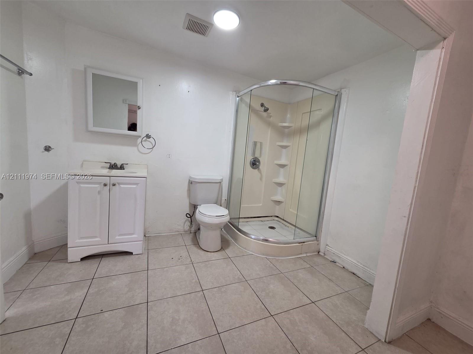 Bathroom featuring tile flooring, a white vanity cabinet with an integrated sink, a mirrored medicine cabinet, a toilet, and a curved glass-enclosed shower stall with built-in shelving