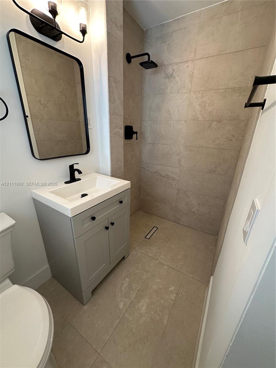 Bathroom featuring a walk-in shower with large-format tile surround, grey vanity with integrated sink, rectangular wall-mounted mirror, and matte black fixtures