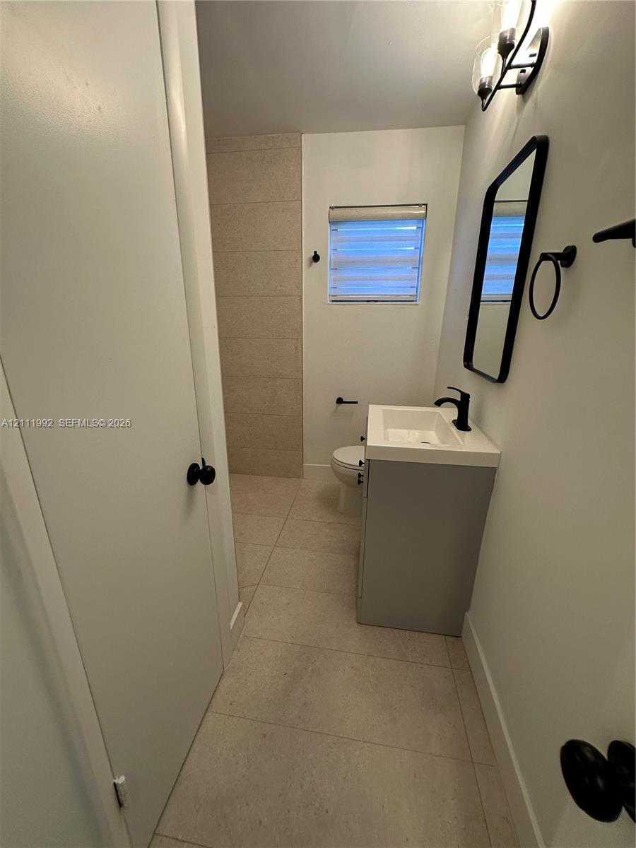 Bathroom featuring large-format neutral tile flooring, a modern grey vanity with a white rectangular sink, matte black hardware, and a framed vanity mirror