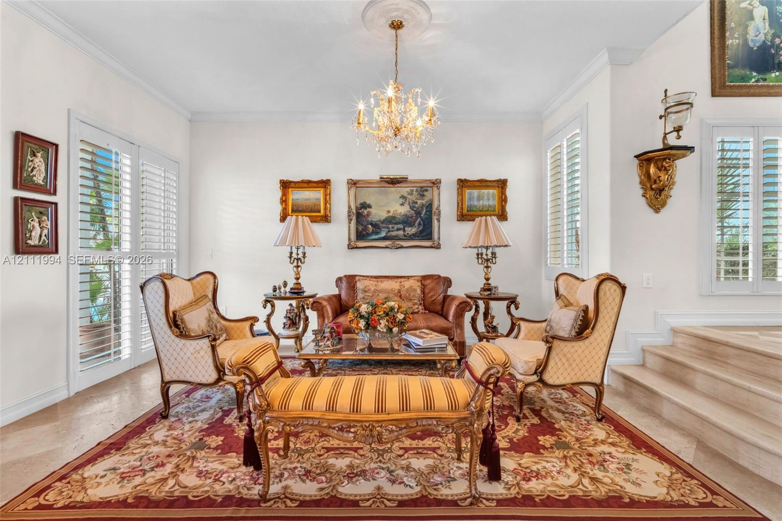 Formal living area featuring polished stone flooring, custom plantation shutters, tiered stone steps, crown molding, and a central crystal chandelier with a decorative ceiling medallion
