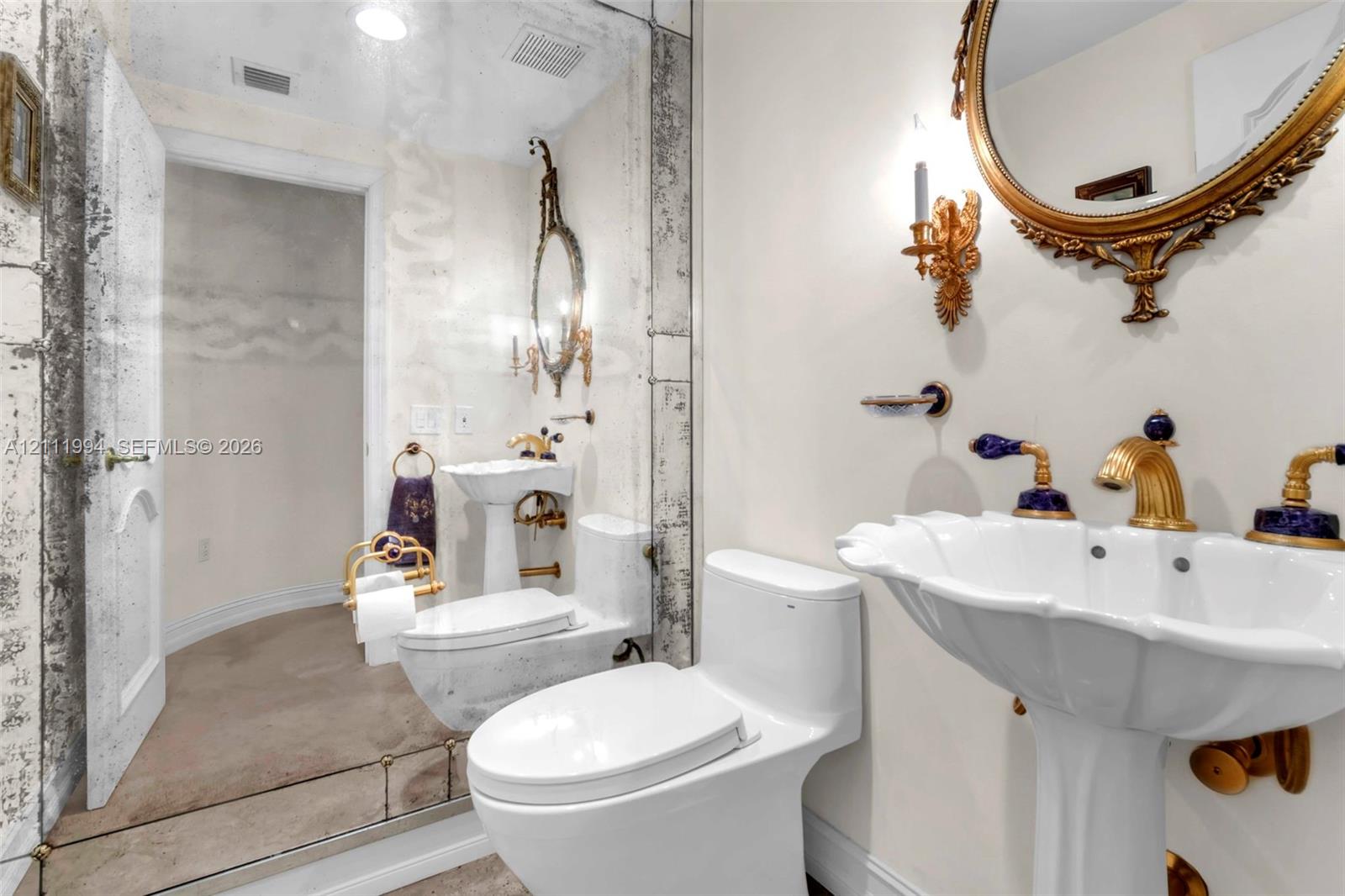 Pedestal sink with scalloped basin, brass-finish faucet with blue accent detailing, mirrored wall paneling, and decorative wall-mounted lighting