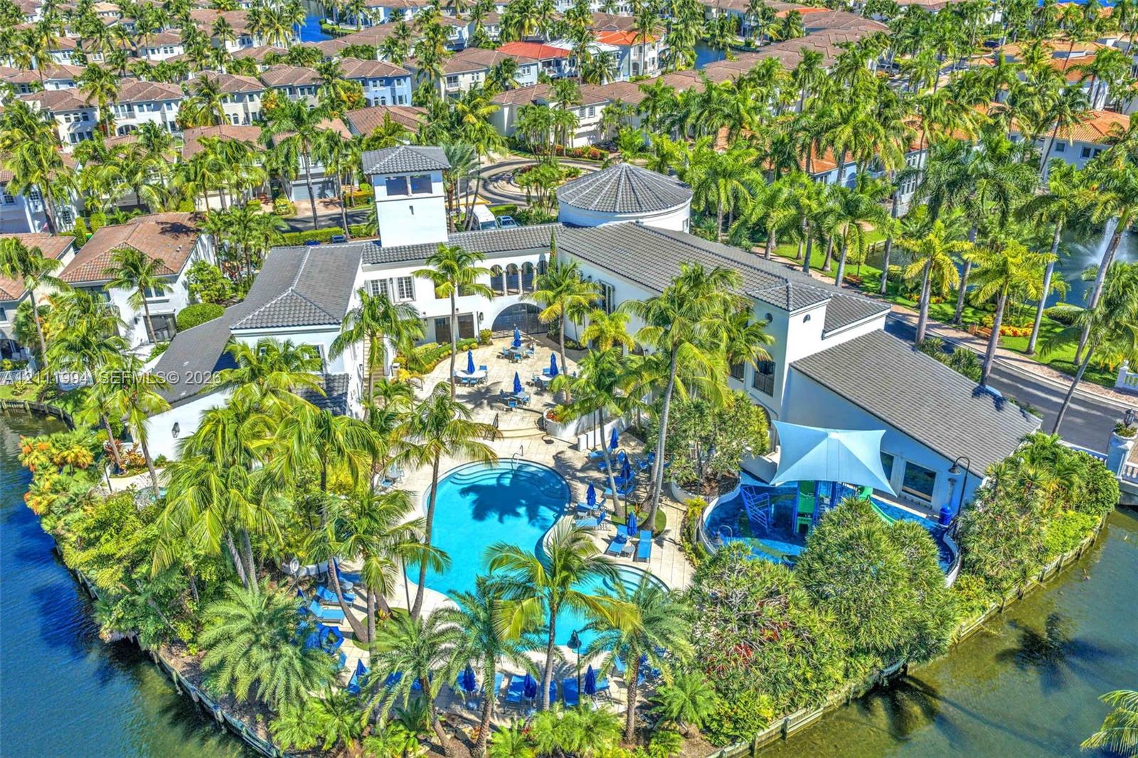 Community clubhouse featuring a tiled roof, architectural tower, swimming pool, water-feature area, and surrounding palm trees