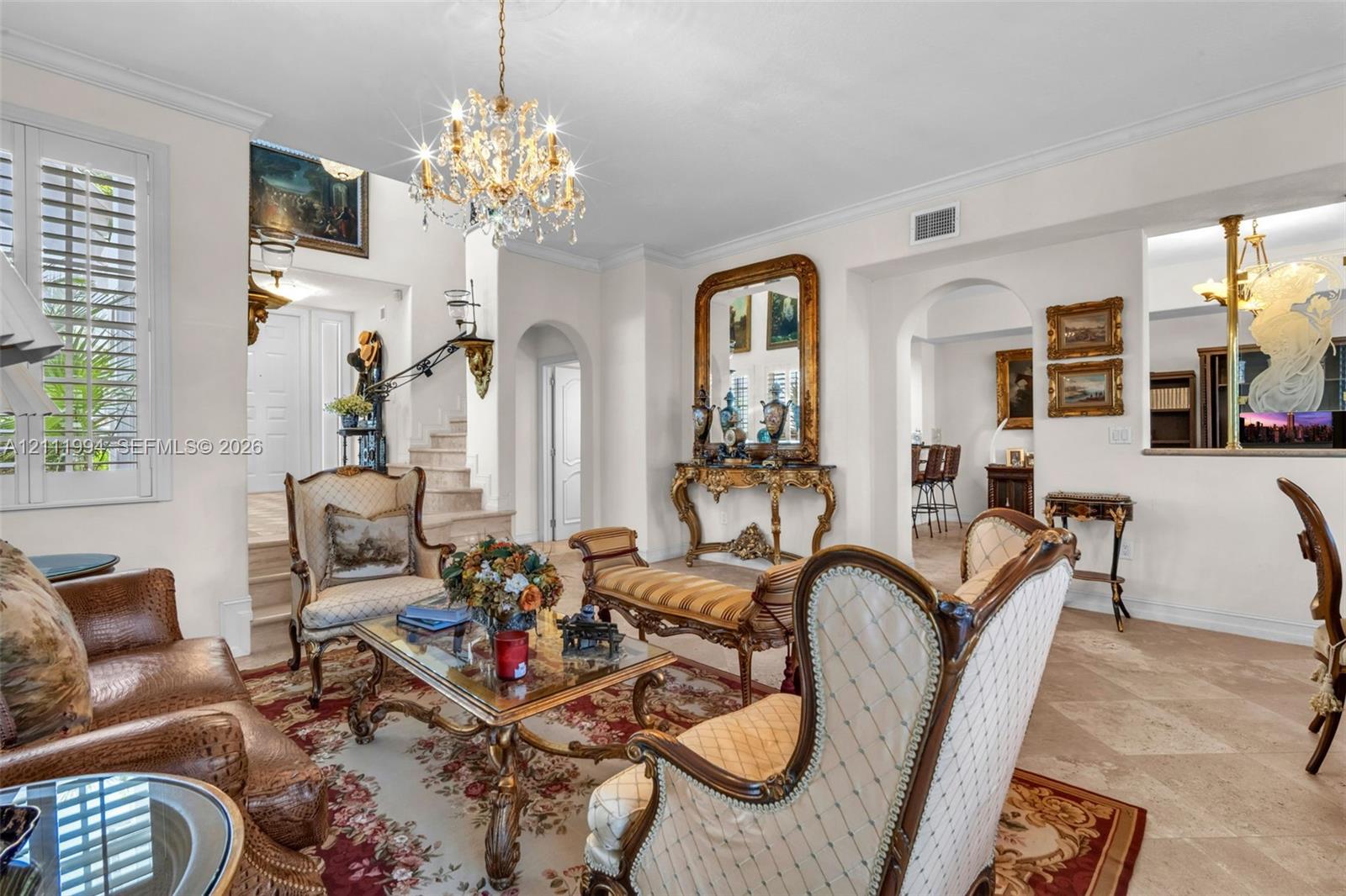Living area featuring stone flooring, ornate crystal chandelier, arched doorways, crown molding, and plantation shutters
