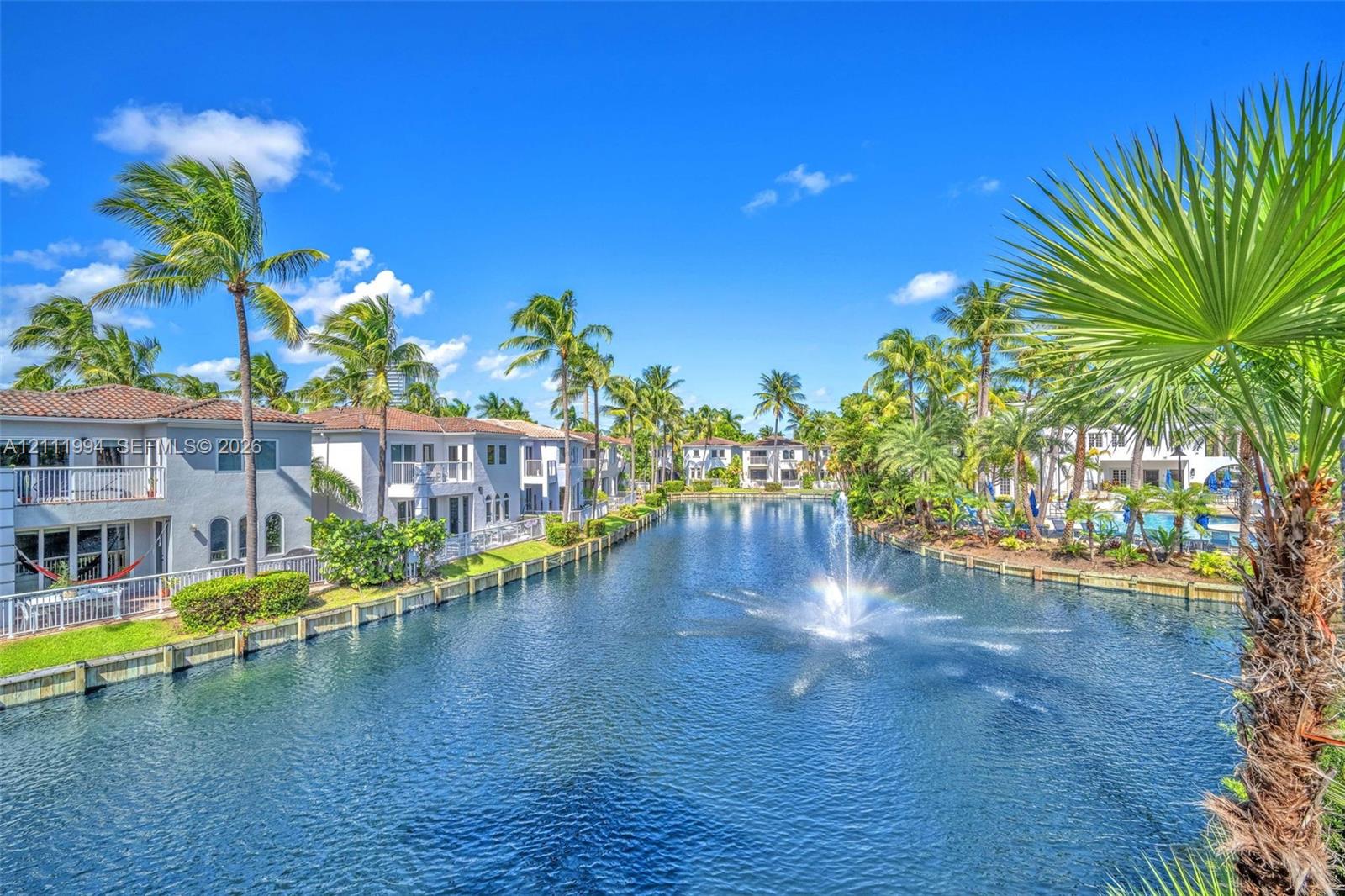 Waterfront complex featuring a central fountain, tile-roofed multi-story structures, concrete bulkheading, and mature palm trees
