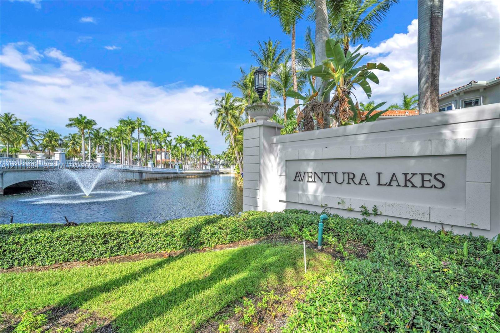 Community entrance featuring a concrete monument sign, water feature, bridge, and mature palm trees