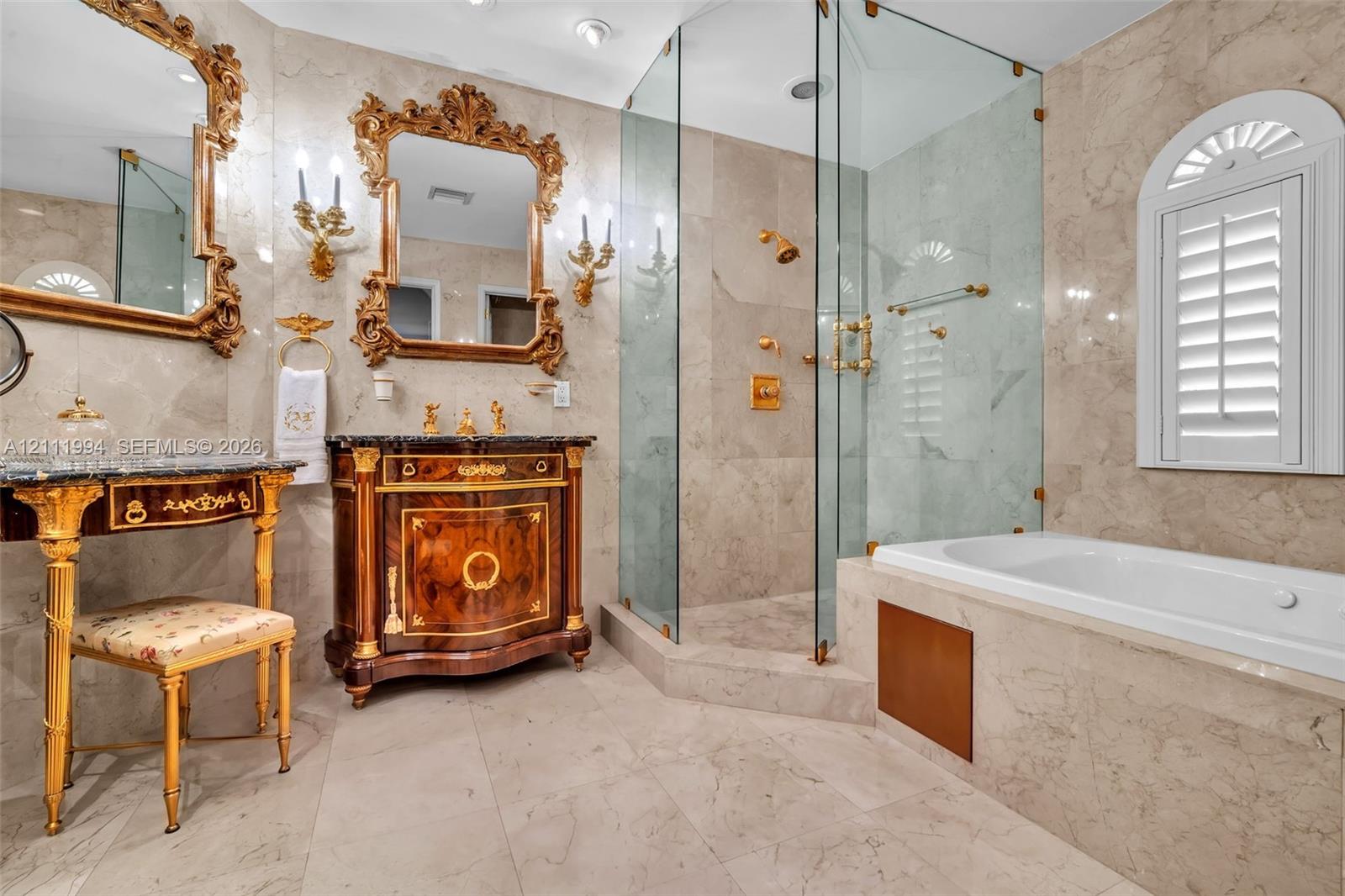 Stone-tiled bathroom featuring a glass-enclosed shower, a built-in soaking tub, a custom vanity with gold-tone accents, and wall-mounted light fixtures
