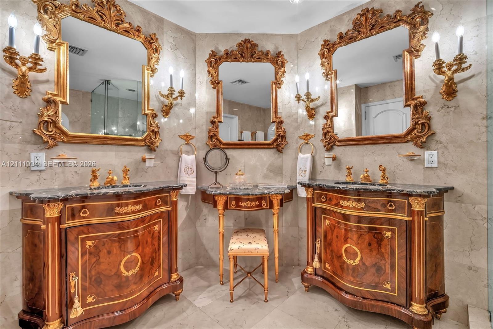 Custom dual vanities with dark stone countertops, ornate gold-leaf framed mirrors, integrated central makeup vanity, gold-tone wall-mounted candle-style sconces, and neutral tile flooring