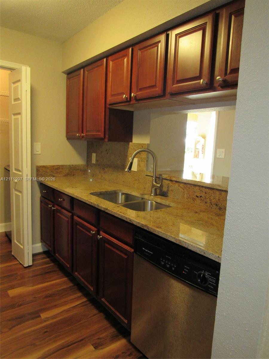 Rich-toned wood cabinetry, stone countertops, stainless steel dual-basin sink with arched faucet, stainless steel dishwasher, and wood-finish flooring