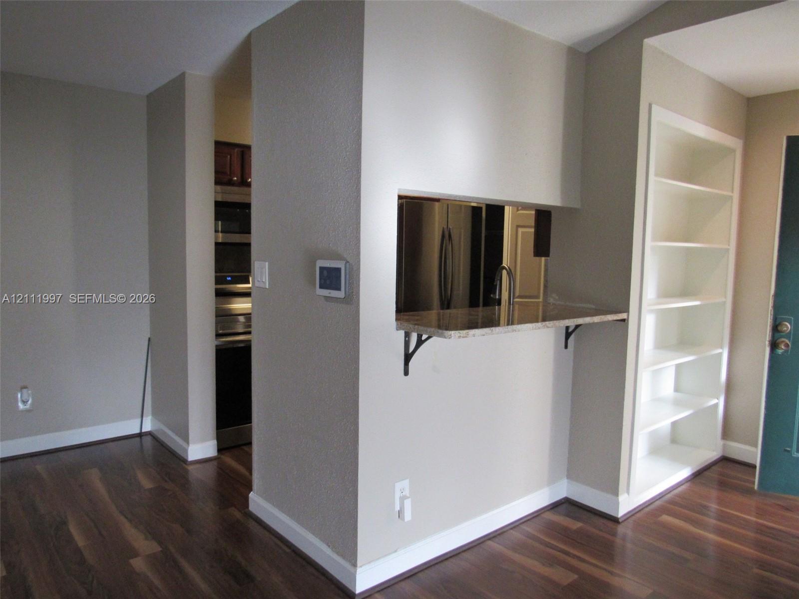 Built-in shelving unit, wood-finish flooring, pass-through opening with a stone countertop, and stainless steel kitchen appliances