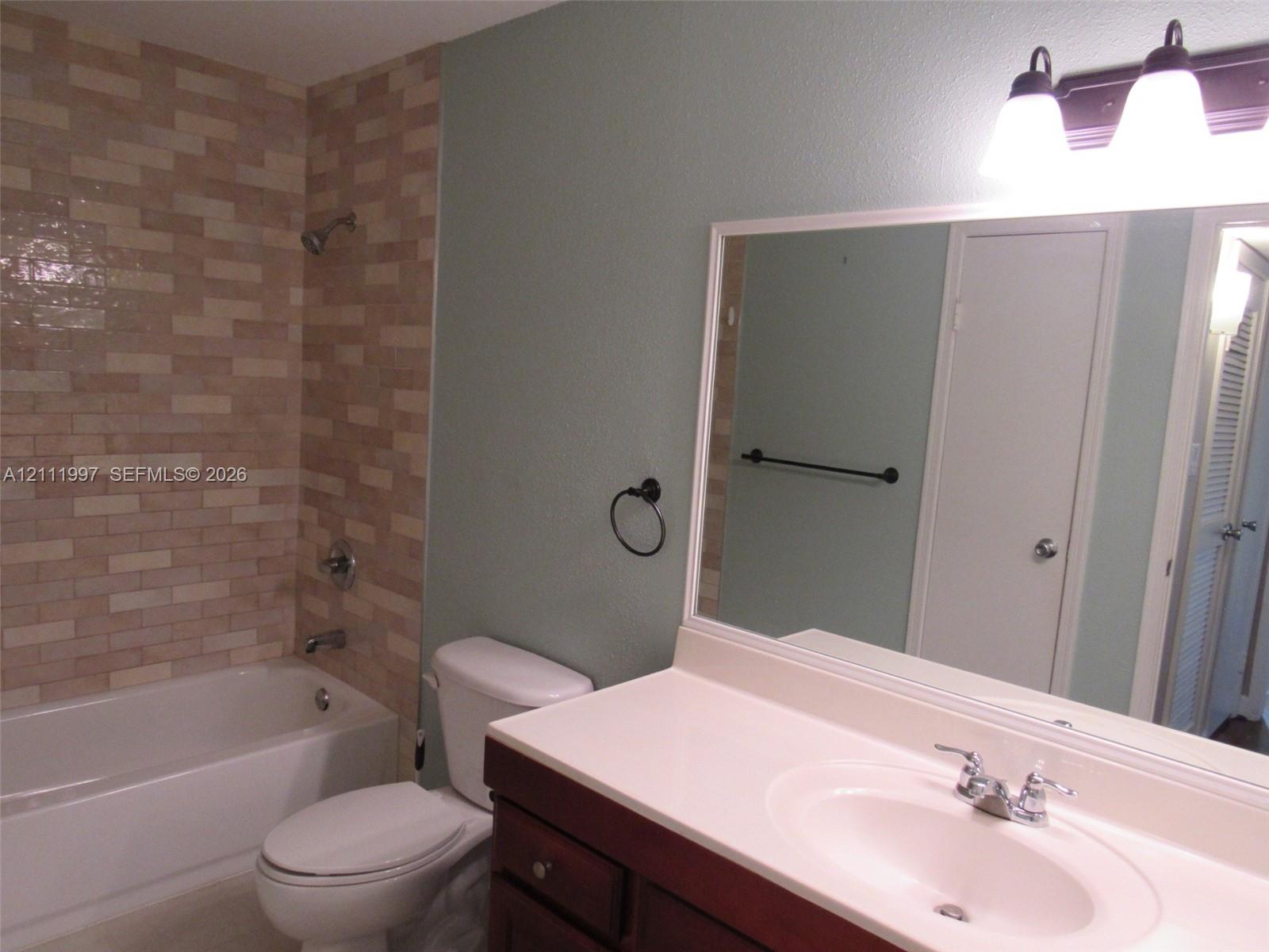 Bathroom featuring a shower and bathtub combination with rectangular tile surround, dark wood vanity, solid-surface countertop with an integrated sink, and triple-bulb wall-mounted vanity lighting