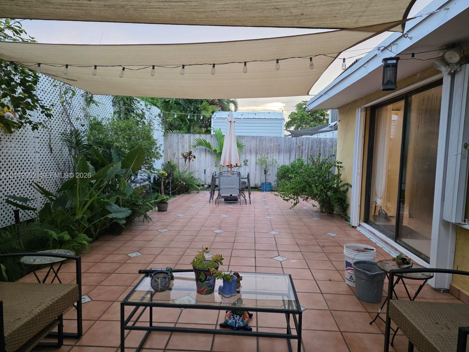 Spacious patio featuring terra-cotta tile flooring with decorative inlay, overhead shade sail, white lattice screening, and sliding glass door access
