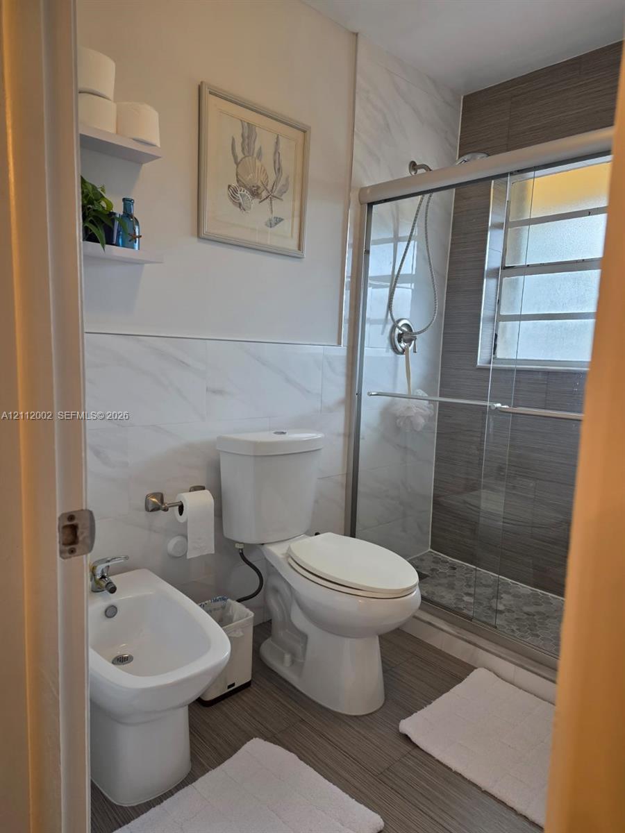 Full bathroom featuring a walk-in shower with glass sliding doors, ceramic wall tiling, a built-in bidet, and two floating wall shelves