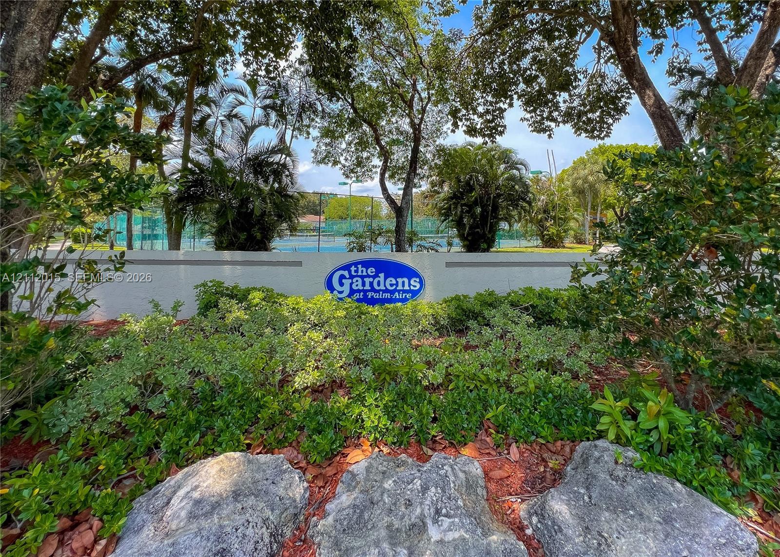 Landscaped entrance featuring mature shade trees, stone boulders, shrubbery, and signage for The Gardens at Palm-Aire