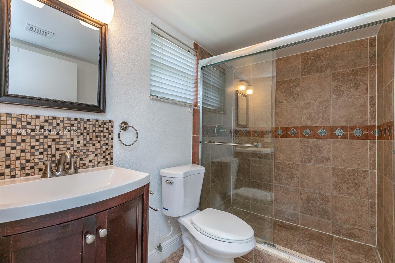 Bathroom featuring a dark wood-finish vanity, decorative mosaic tile backsplash, framed wall mirror, and walk-in shower with glass enclosure and tiled surround