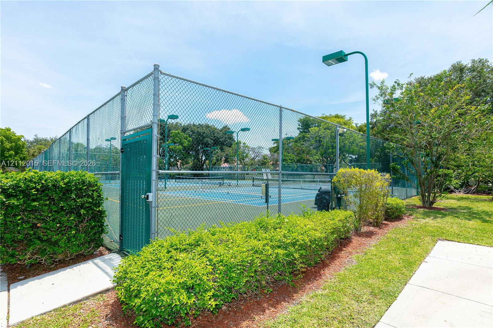 Tennis court enclosed by chain-link fencing, featuring overhead lighting, a gated entrance, and manicured landscaping