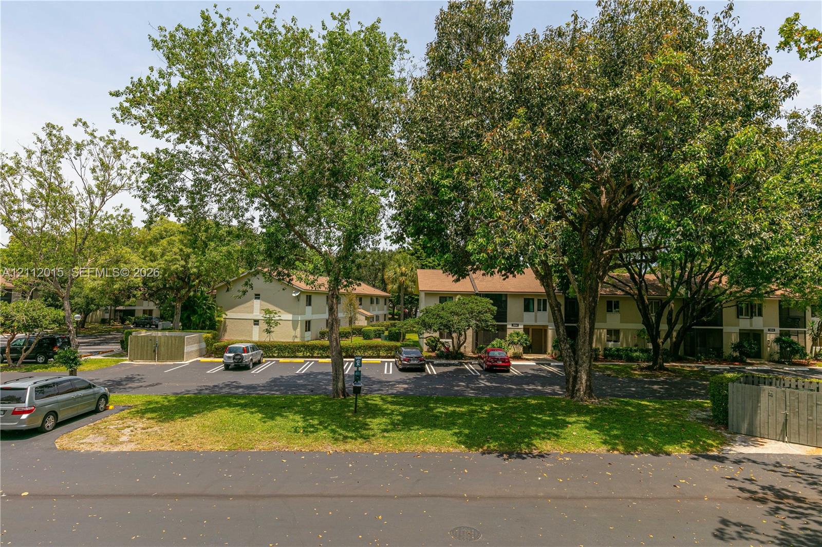 Exterior view featuring multi-story residential buildings, paved surface parking, mature trees, and landscaped shrubbery