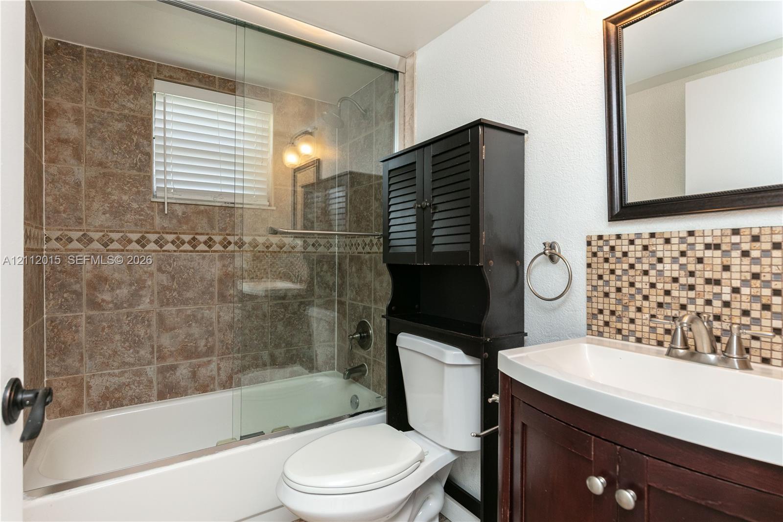 Bathroom featuring a shower-tub combination with glass enclosure, tiled surround with decorative accent border, mosaic tile backsplash, dark wood-finish vanity, and wall-mounted mirror