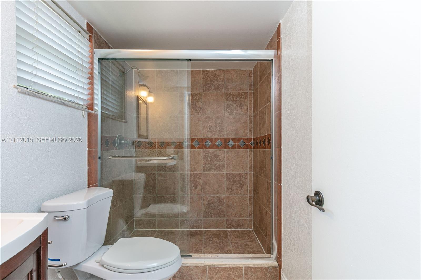 Walk-in shower featuring glass sliding doors and decorative tile inlay, ceramic tile wall finishes, and a neutral-tone commode