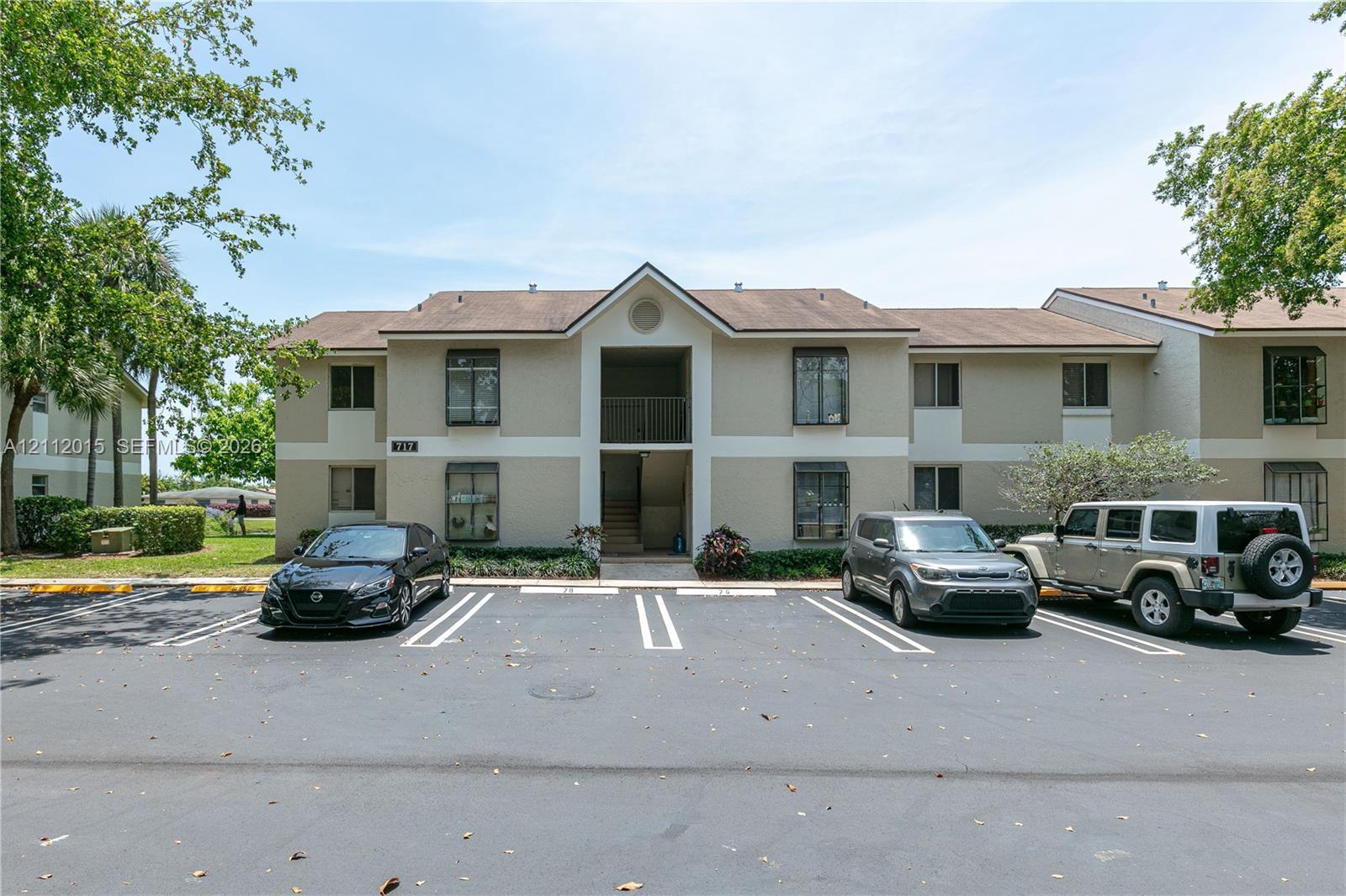 Multi-story building exterior featuring a neutral stucco facade, shingle roof, dark-frame windows with metal awnings, and a ground-level parking area
