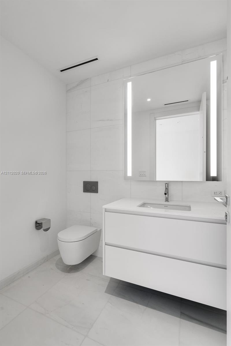 Modern floating vanity featuring a smooth-surface countertop, wall-mounted commode, large integrated LED-lit mirror, and large-format wall and floor tiles