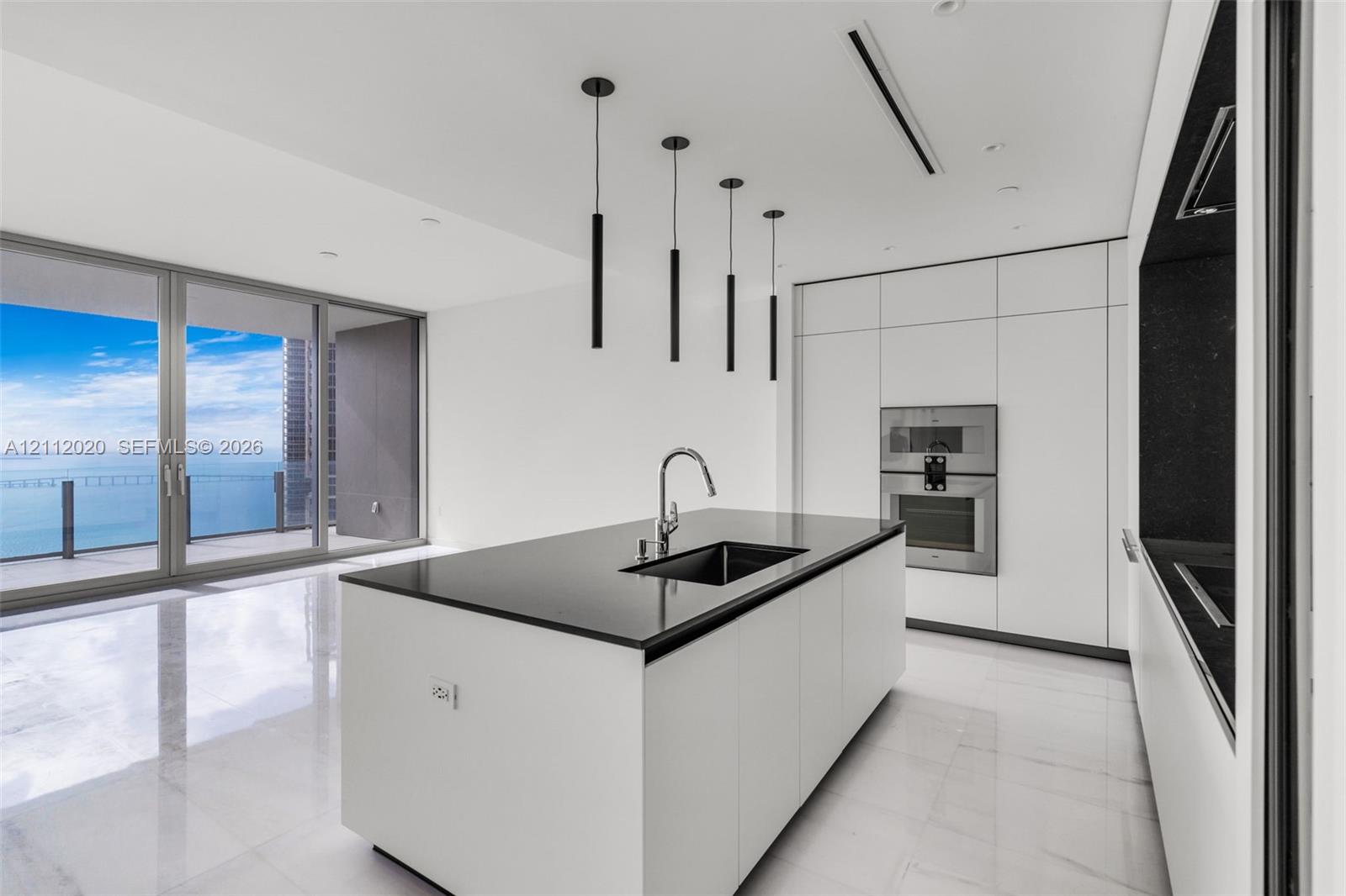 Modern kitchen featuring a central island with a stone-surface countertop, integrated wall-mounted double ovens, minimalist flat-panel cabinetry, stone flooring, and linear pendant lighting