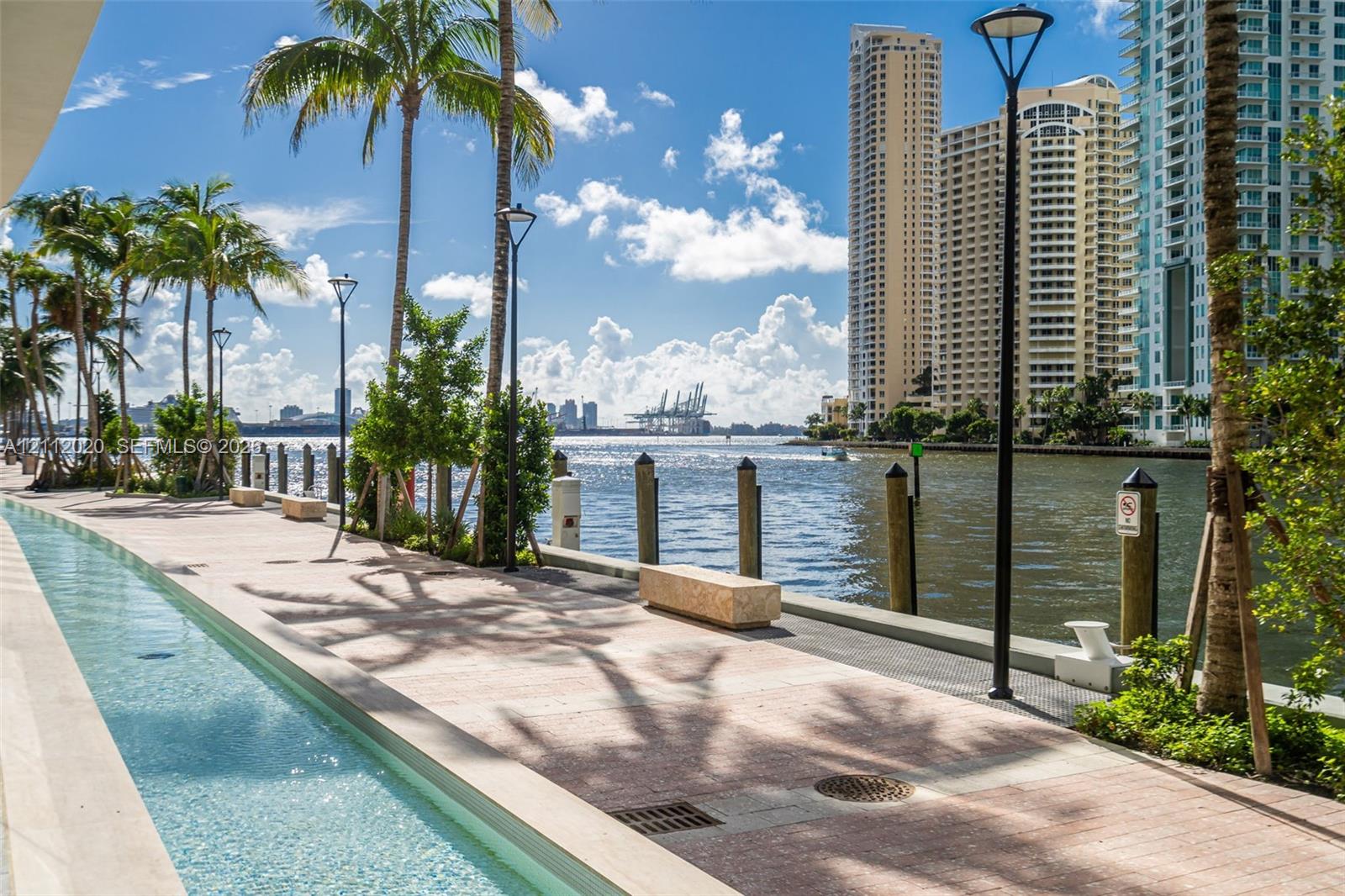 Waterfront promenade featuring a tiled walkway, a decorative linear water feature, integrated stone benches, and mature palm trees