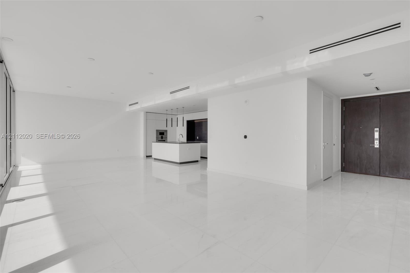 Open-concept interior featuring large-format tile flooring, recessed ceiling lighting, linear air diffusers, a kitchen island with built-in cabinetry, and a dark-finish double-door entry Open-concept interior featuring large-format tile flooring, recessed ceiling lighting, linear air diffusers, a kitchen island with built-in cabinetry, and a dark-finish double-door entry