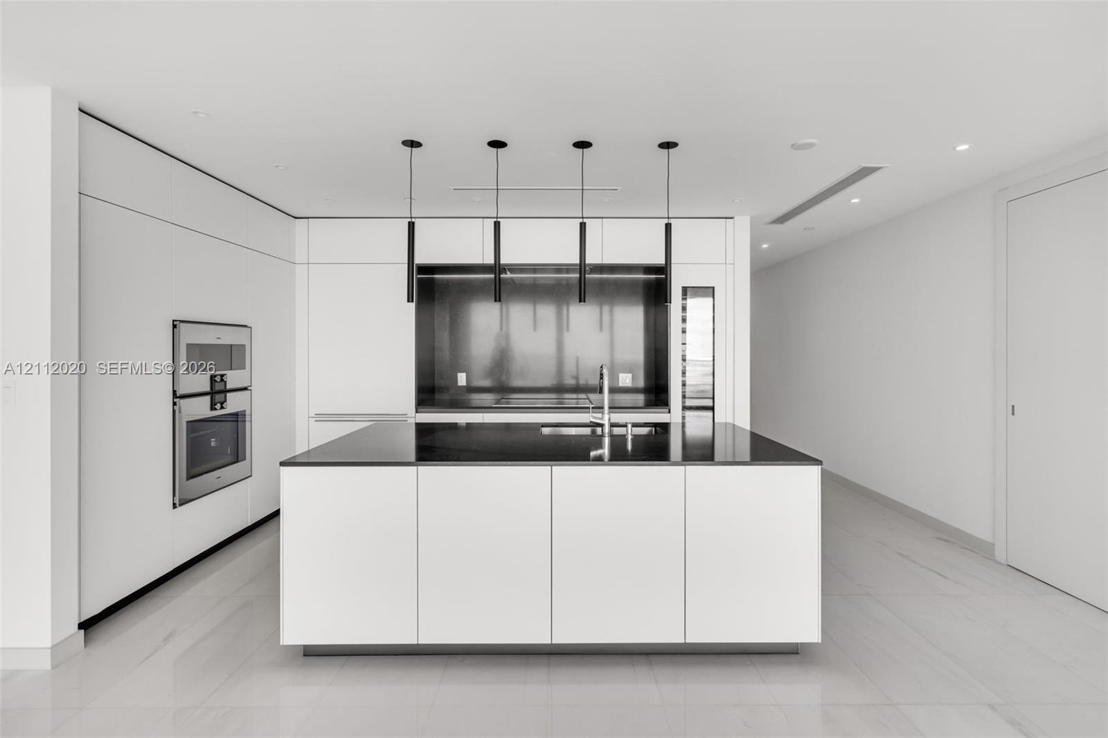 Minimalist kitchen featuring flat-panel cabinetry, a central island with a contrasting stone countertop, built-in stainless steel double ovens, a metal backsplash, and suspended cylindrical pendant lighting