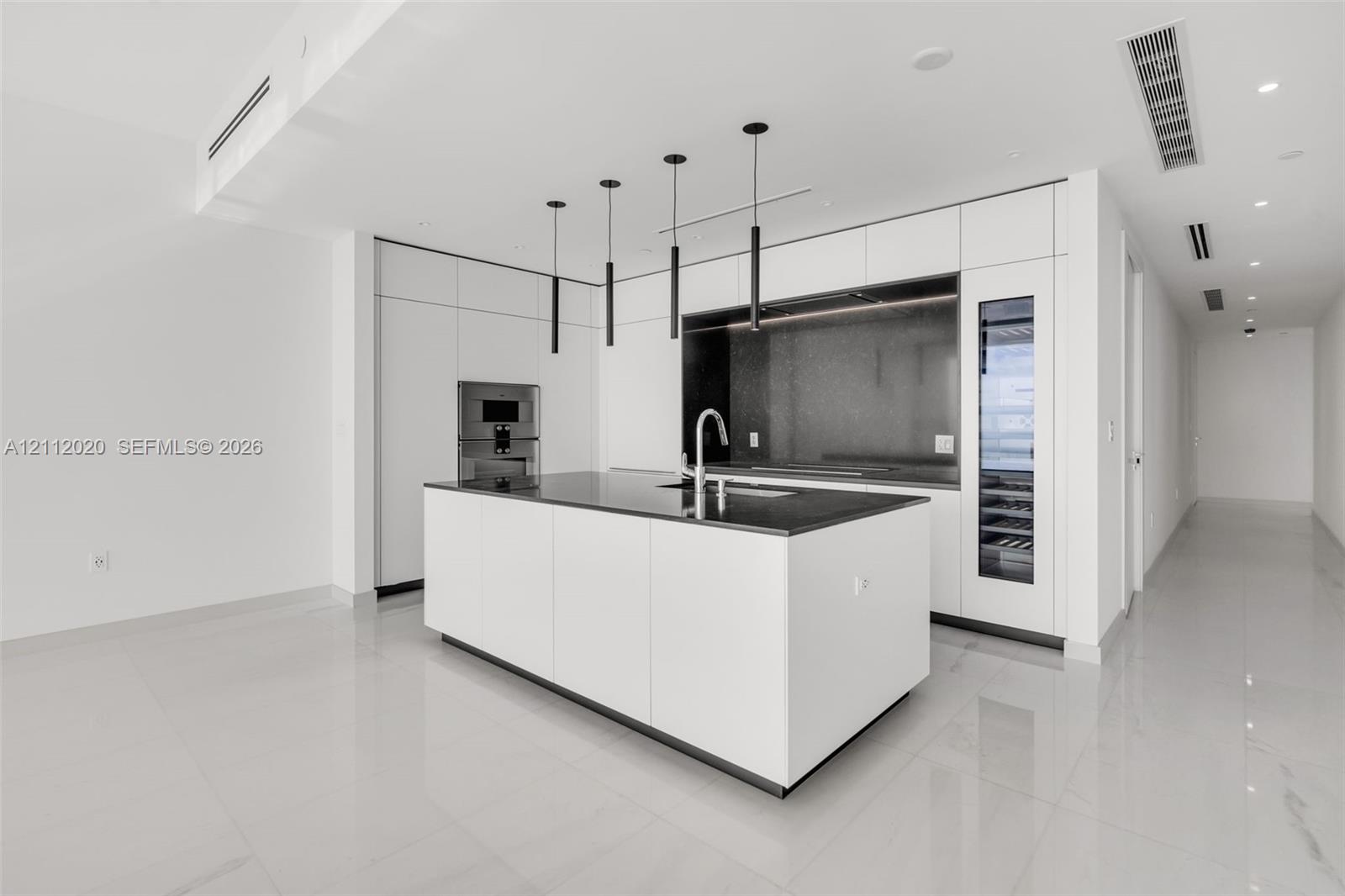 Modern kitchen featuring white flat-panel cabinetry, a central island with a dark countertop, integrated wine refrigeration, minimalist pendant lighting, and polished stone flooring