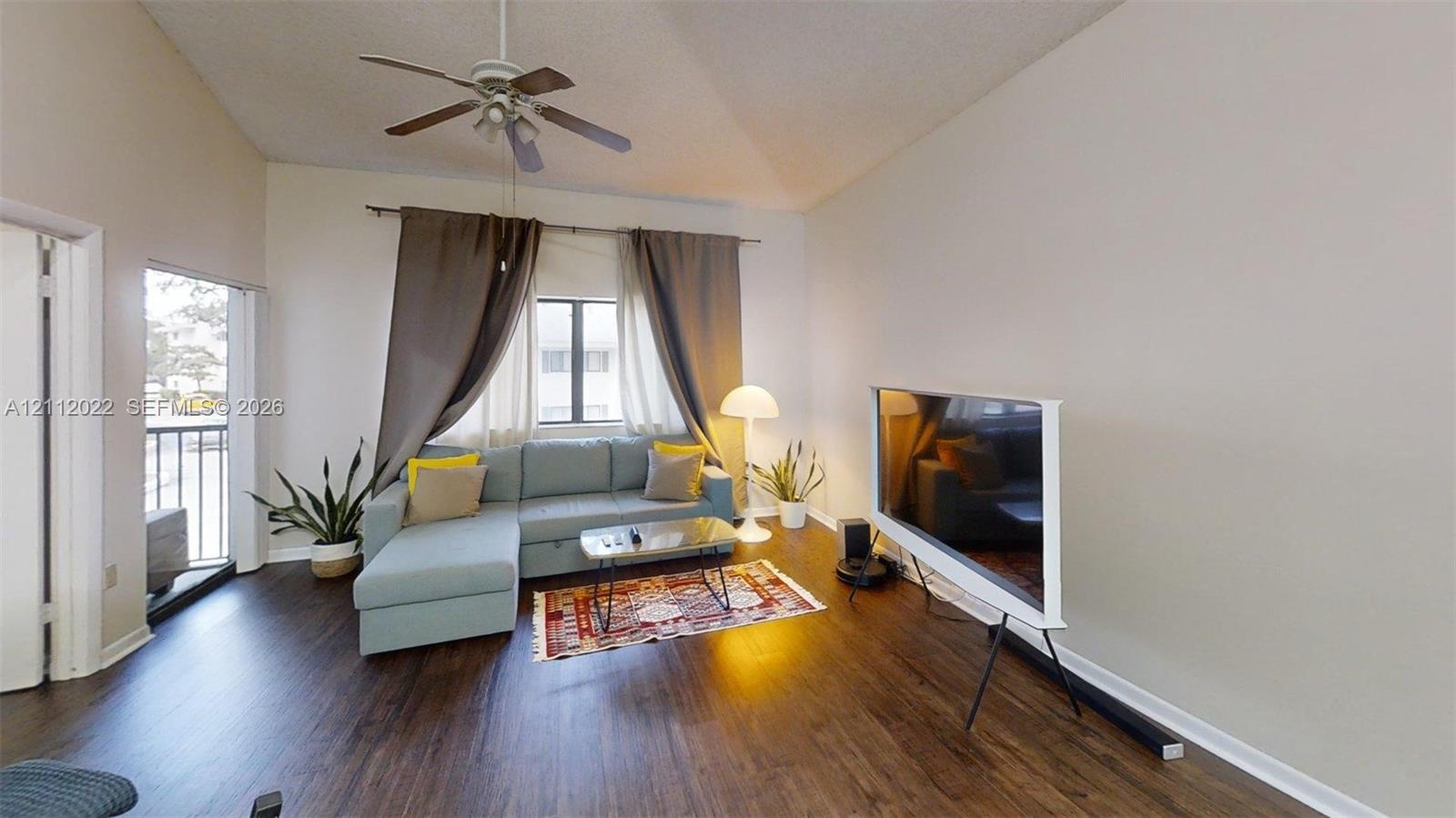 Living area featuring vaulted ceilings, wood-finish flooring, a ceiling fan with integrated lighting, and a private balcony access point